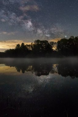 Gece vakti, kıyı şeridinde ağaçların silueti olan küçük bir gölün üzerinde Samanyolu