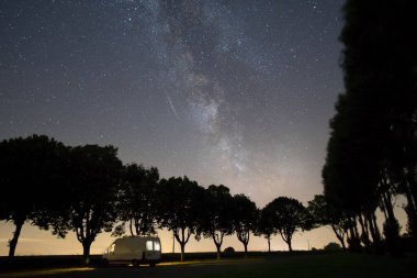 Milky Way over french countryside with camper van and silhouette of trees at night, Normandy, France