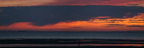 Panorama of colorful dramatic red sky with clouds in the twilight after sunset over the ocean with silhouette of child running at the beach