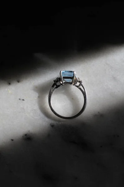 A side view of a silver ring with a big blue stone laying on marble with dramatic lighting.