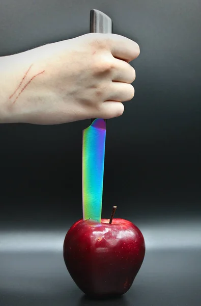 A human hand with scratches holding a multi-colored kitchen knife stabbing a red apple with a black background.