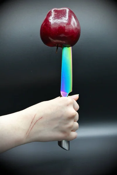 An apple upside down on the point of a rainbow colored knife held upside down by a scratched hand with a black backgrounds.