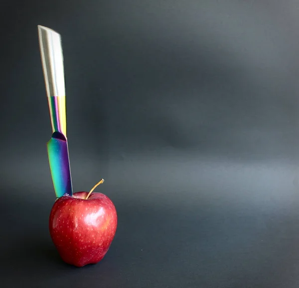 A rainbow colored knife stabbed into a red apple with a black background.