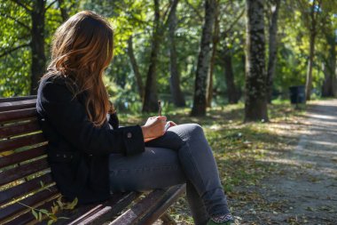 Konsantre olmuş bir kadın parkta bankta oturmuş deftere yazı yazıyor ya da çizim yapıyor..