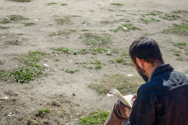 Kulaklıkla derin derin düşünürken parkta kitap yazan ya da çizen bir adam. Arkadan görüntüle.