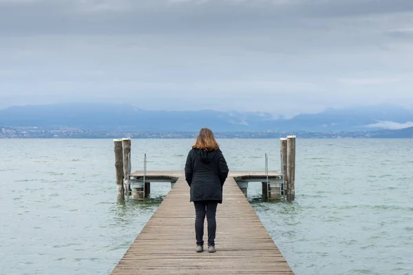 Woman standing in the middle of a pier on the Lake di Garda, italy, with snow capped mountains and clouds in the background. 
