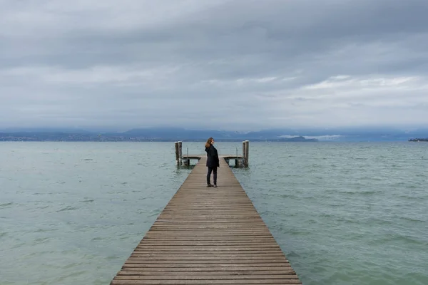 Woman standing in the middle of a pier on the Lake di Garda, italy, with snow capped mountains and clouds in the background. 