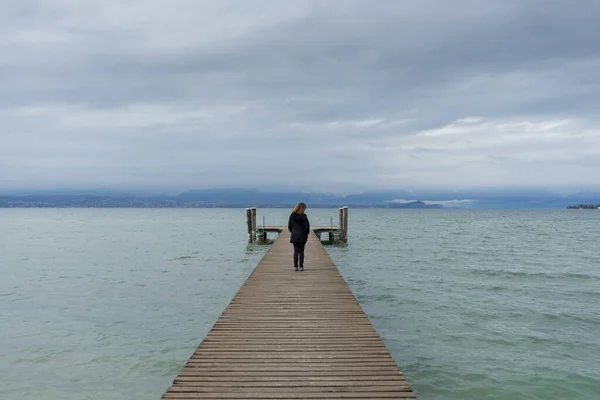 Woman standing in the middle of a pier on the Lake di Garda, italy, with snow capped mountains and clouds in the background. 