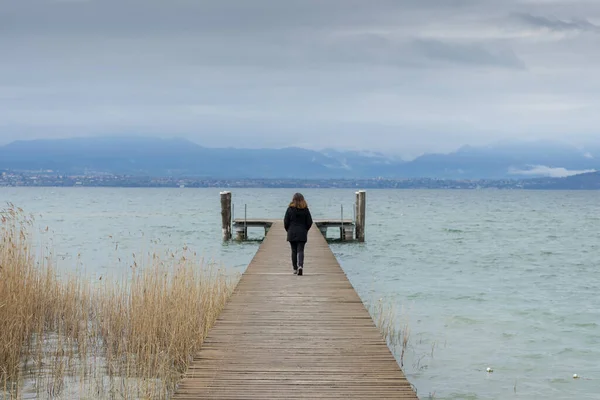 Woman standing in the middle of a pier on the Lake di Garda, italy, with snow capped mountains and clouds in the background. 