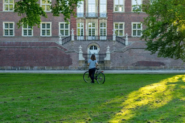 Young woman next to her bicycle in the middle of a park at sunset, with a historical building in the background 