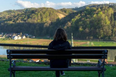 Slovenya köyüne tepeden bakan ahşap bankta oturan bir kadın, arkasında nehir ve dağlar var. Yüksek kalite fotoğraf
