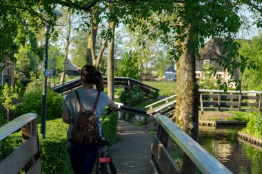 Sırtı tahta bir köprüde dikilen genç bir kadın. Yanında bisikleti, etrafı ağaçlarla çevrili ve yanında bir nehir var. Yüksek kalite fotoğraf