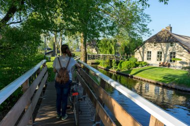 Sırtı tahta bir köprüde dikilen genç bir kadın. Yanında bisikleti, etrafı ağaçlarla çevrili ve yanında bir nehir var. Yüksek kalite fotoğraf