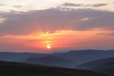 Çayırların ve ormanların üzerinde gün doğumu. Ormanlar ince bir sis içinde. Gökyüzünde bulutlar ve güneş var, bu da yumuşak bir güneş yaratıyor..