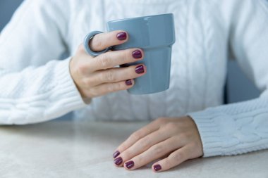 Beyaz kazaklı, elinde gri bir kupayla tanınmayan bir kadın bir eli masada dinleniyor, tırnakları boyalı..