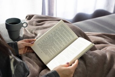 Bir kadının elleri yanında bir fincan kahveyle kitap tutuyor..