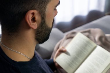 Sakallı genç adam evde kitap okurken kanepede oturuyor.