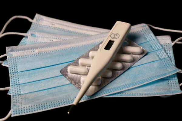 İlaç tabletinin üstündeki dijital termometre ve siyah arkaplandaki mavi maskeler.