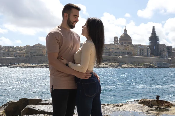 Birbirine aşık çiftler birbirlerine bakar ve arka planda La Valleta, Malta ile deniz kıyısında kucaklaşırlar..