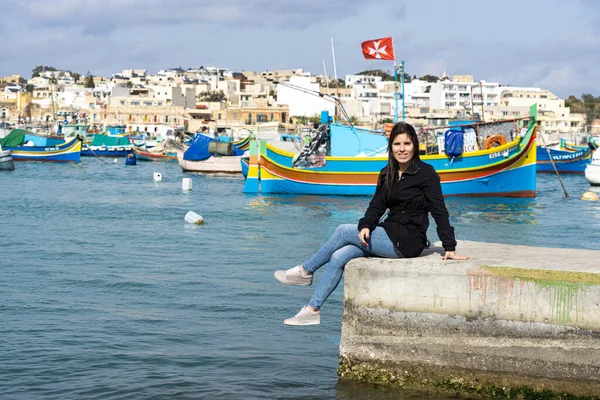 Genç bayan turist oturuyor ve arka planda Marsaxlokk, Malta 'da renkli teknelerle dolu bir deniz var..