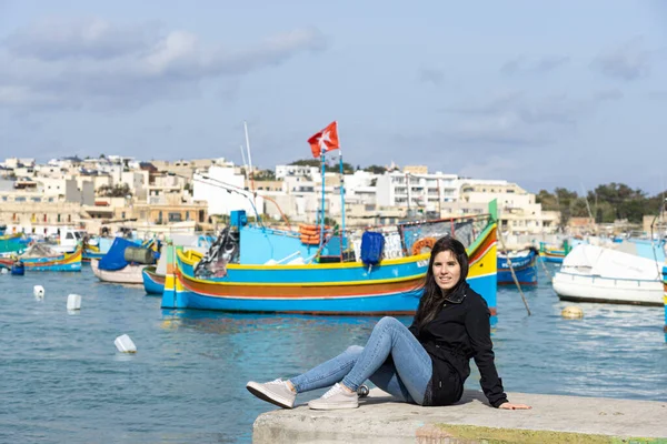 Genç bayan turist oturuyor ve arka planda Marsaxlokk, Malta 'da renkli teknelerle dolu bir deniz var..