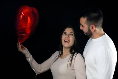 Young couple happy and smiling while holding a red heart shaped balloon on valentine's day with a black background.