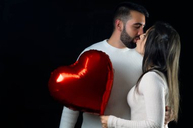 Young couple in love holding a red heart-shaped balloon while kissing each other.