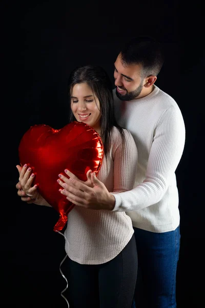 Happy young couple hugging a heart shaped balloon in foreground with black background.