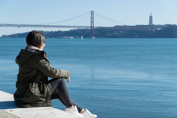 Arka planda 25 Nisan köprüsü olan genç bir kadın oturuyor, Lisbon, Portekiz