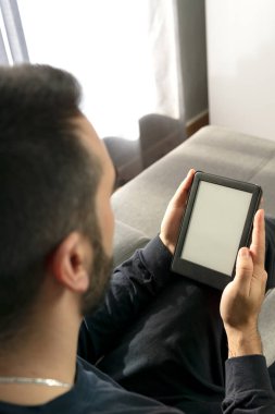 Evde rahatlatıcı bir anda elinde bir fincan kahve tutarken elektronik kitapla kitap okuyan tanınmamış bir genç adam..