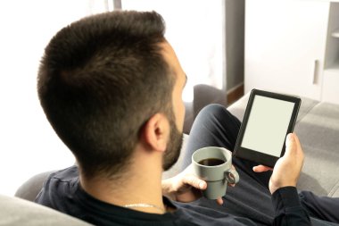 Odaklanamayan genç adam e-kitabıyla okuyor ve evdeki kanepesinde bir fincan kahve tutuyor..