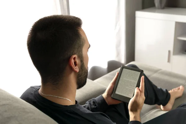 Evdeki kanepede otururken elektronik kitabıyla kitap okuyan tanınmamış genç bir çocuk..