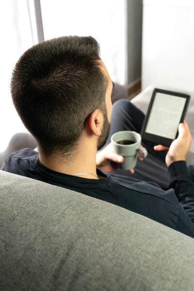 Unrecognizable young boy photographed from behind while reading with his electronic book and holding a cup of coffee in a moment of relaxation and disconnection.