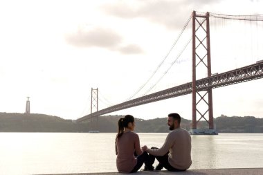 25 Nisan Köprüsü ve önünde bir çiftle birlikte Tagus Nehri 'nin kıyısında el sıkışan Kral İsa heykeli..