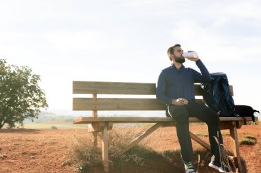 Sakallı adam, tahta bir bankta dinlenirken şişesinden su içiyor. Spor ve sağlık konsepti.