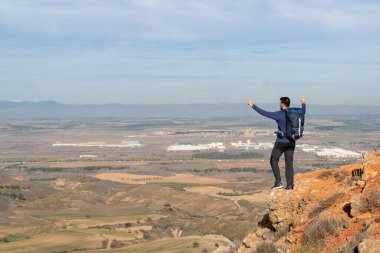 Sırtında bir sırt çantasıyla uzaktaki şehre bakarken kollarını kaldıran genç sakallı çocuk. Başarı ve kendini geliştirme kavramı..