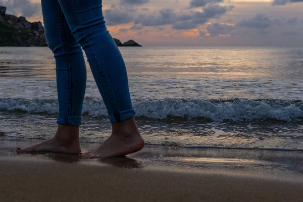 Detail of the legs of an unrecognizable woman walking on the beach with the sea, rocks and sunset in the background.