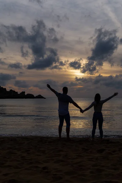 Unrecognizable couple in love raising their hands on the shore of the beach at sunset.