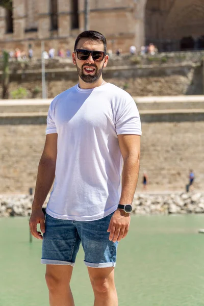 Young man wearing sunglasses, denim shorts and a white t-shirt posing with the background out of focus.