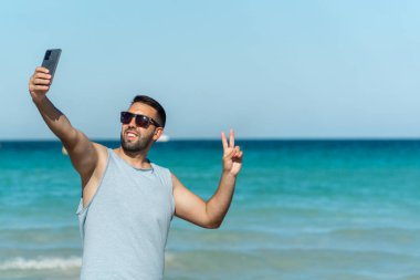 Young man with beard and sunglasses taking a selfie with his smartphone on the beach on a sunny day with plenty of copy space.