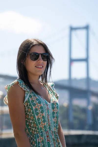 Ön planda güneş gözlüklü bir kadının portresi ve arka planda odaktan uzak ünlü İstanbul köprüsü..
