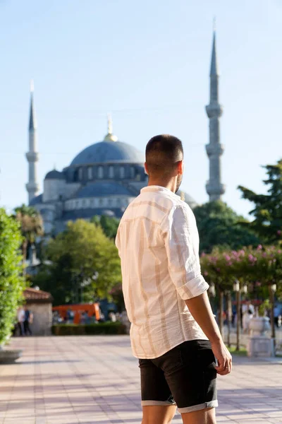 Sırtüstü yatmış, gün batımında arka planda mavi camiyle poz veren tanınmamış genç adam..