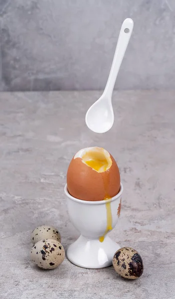 Boiled soft-boiled egg in egg cup with spoon, quail eggs on concrete background