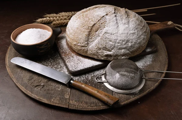 A round loaf of freshly baked sourdough bread flavored with hemp flour, with a knife on a cutting board. Craft bread with seeds on a dark table. Country style sourdough bread