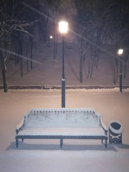 提灯を背景にした夜の公園の冬の雪に覆われたベンチ — ストック写真