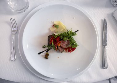 Bir tabak az pişmiş biftek, roket, vişneli domates ve bir restorandaki beyaz masada parmesan.