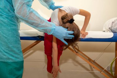 Bir fizyoterapist ortopedik yatakta başı ve vücudu öne eğilmiş bir kız için rehabilitasyon terapisi uygular. Kız sağ koluyla sol ayağına dokunuyor..