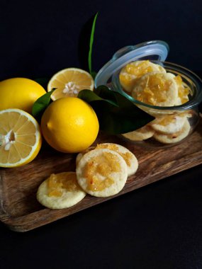Lemons fresh whole and cut with leaves. Cookies with pieces of carmelized lemons on a wooden tray and in a glass jar. Black background.There is a place for an inscription.
