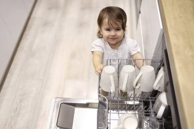 Little cute toddler girl helping to unload dishwasher. Funny little mommys helper in white kitchen at home. Healthy kid is doing household chores. Child indulges in the kitchen.