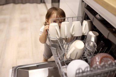 Little cute toddler girl helping to unload dishwasher. Funny little mommys helper in white kitchen at home. Healthy kid is doing household chores. Child indulges in the kitchen.